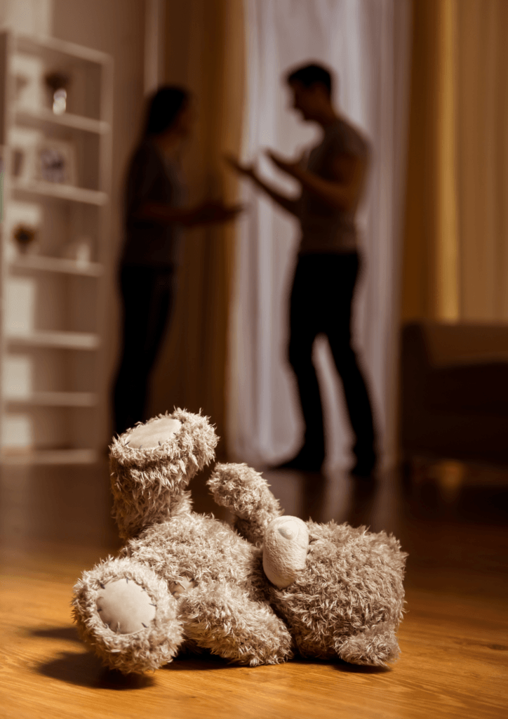 Photo d'un ours en peluche tombé sur le sol avec au loin les parents qui se disputent